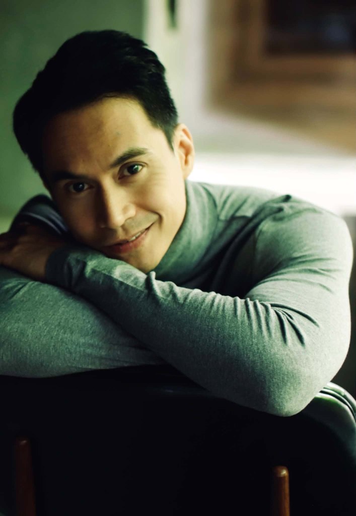 A close-up portrait of a man with short hair, smiling while resting his arms on a chair. He is wearing a gray turtleneck sweater, with a soft, blurred background.