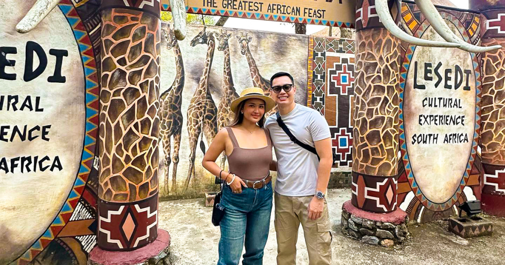A couple stands in front of the Lesedi Cultural Experience entrance featuring vibrant murals of giraffes and decorative elements symbolizing African culture.