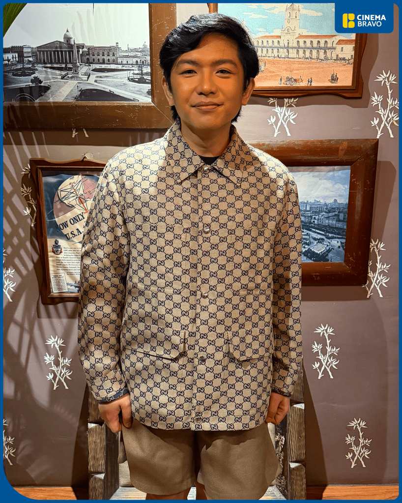 A person wearing a patterned Gucci shirt with shorts, standing in front of framed historical images on a wall.