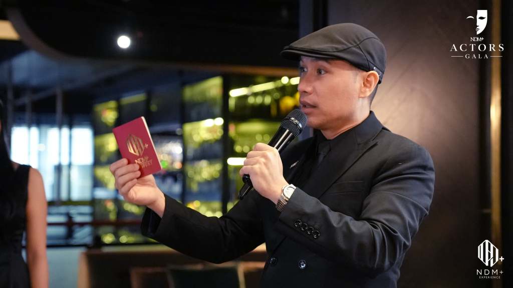 A man in a black suit and cap is speaking into a microphone while holding a red document with a logo. A woman in a black dress is standing nearby, with a blurred background of a modern venue.