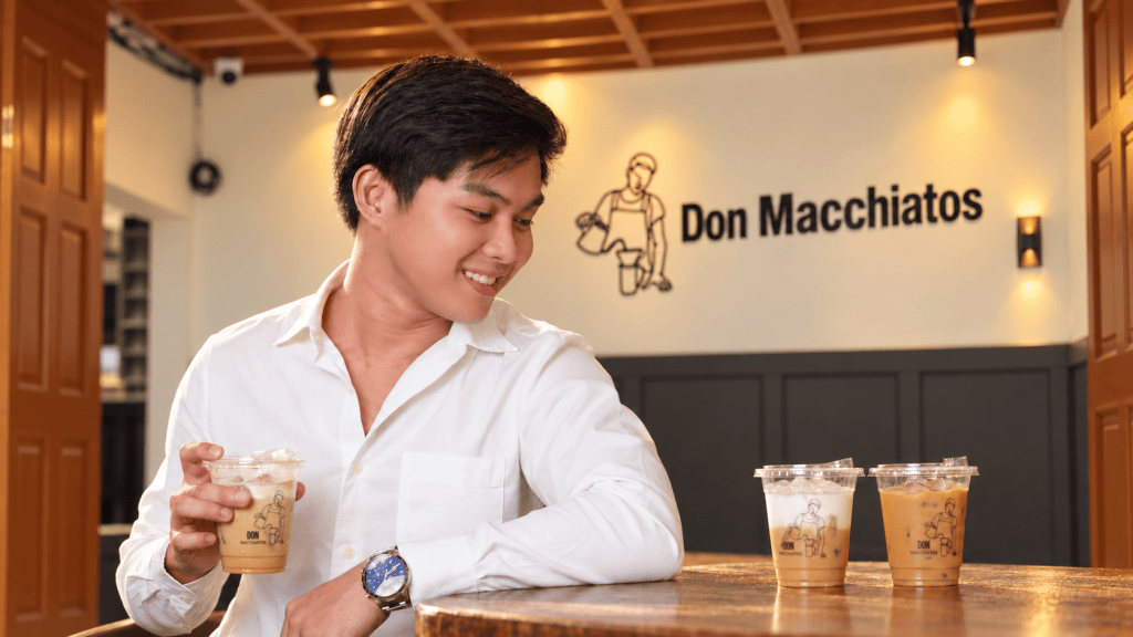 A young man in a white shirt smiles while holding a drink in a coffee shop with the sign 'Don Macchiatos' in the background. Three iced coffee drinks are placed on the table.