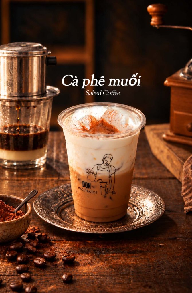 A glass of salted coffee topped with whipped cream and cocoa powder, placed on a decorative plate with a spoon and scattered coffee beans in the foreground, alongside a traditional drip coffee maker.