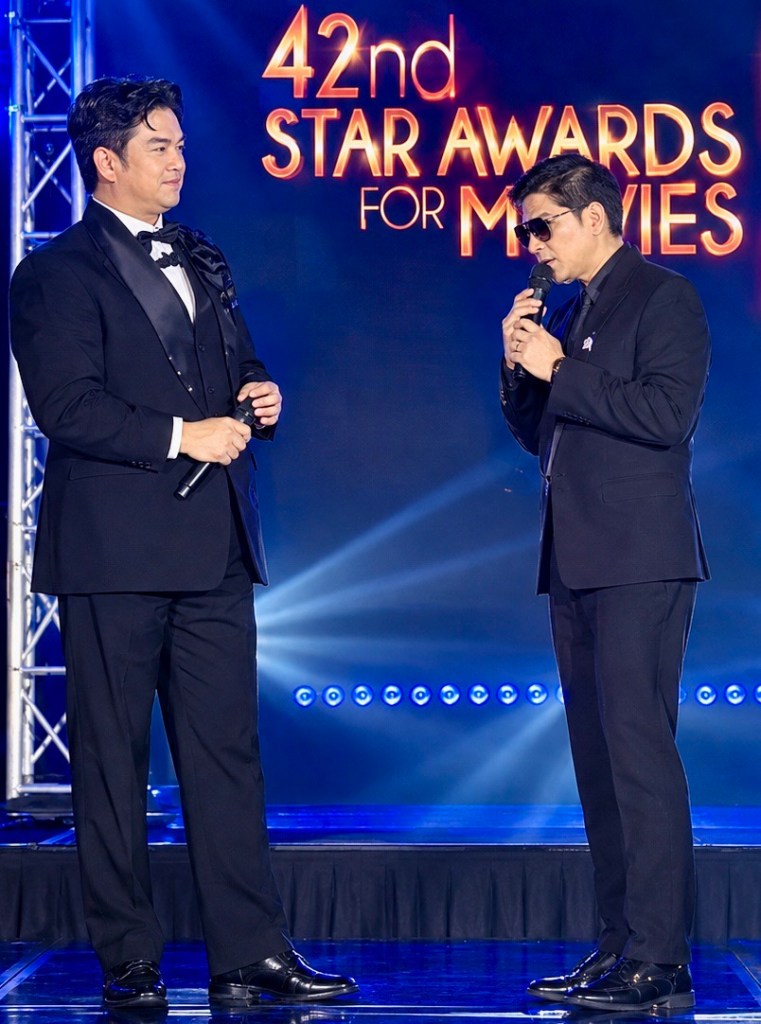 Two male presenters on stage at the 42nd Star Awards for Movies, one holding a microphone and wearing sunglasses, while the other is dressed in a tuxedo.