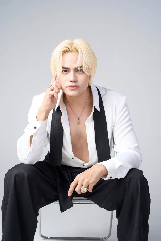 A stylish young man with blonde hair wearing an unbuttoned white shirt and black pants, sitting on a chair and posing thoughtfully.