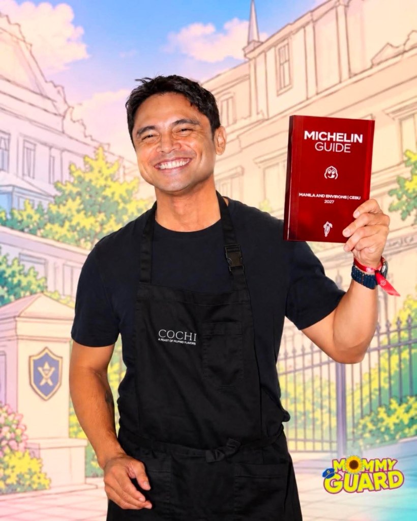A smiling man wearing a black apron holds a Michelin Guide book while standing in front of an illustrated backdrop featuring a building and greenery.